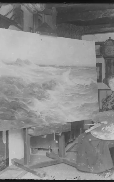 Zwart-witfoto van kunstschilder Betzy Akersloot-Berg zittend op een stoel in haar atelier, voor een groot schilderij van een woeste zee met branding.