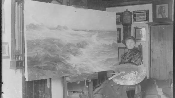 Zwart-witfoto van kunstschilder Betzy Akersloot-Berg zittend op een stoel in haar atelier, voor een groot schilderij van een woeste zee met branding.