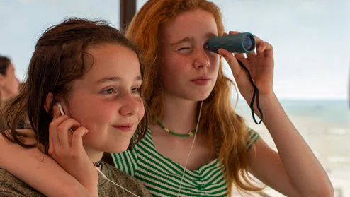 Zusjes kijken met een verrekijker het panorama terwijl ze samen via oortjes naar een audiotour luisteren.